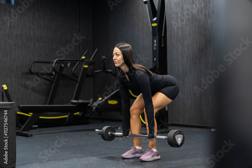 In a sleek gym area, a woman is captured lifting a barbell, her concentration and technique highlighted amidst modern gym structures, blending strength and focus.