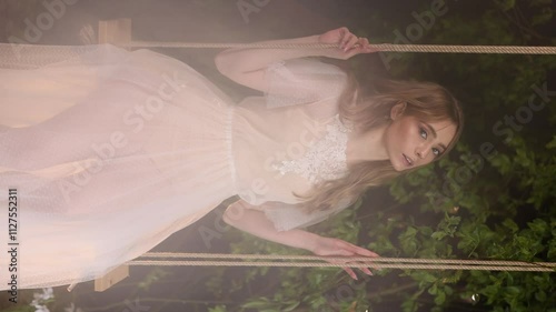 Caucasian woman with long hair in elegant white lace dress sits on wooden swing among dark foliage. Natural cosmetics or perfume concept.
