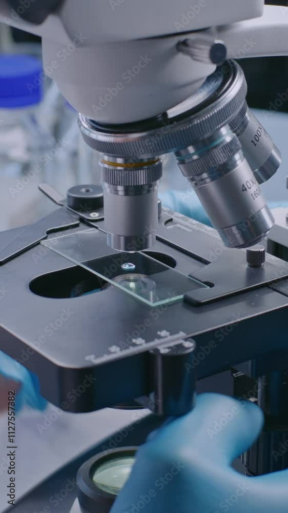 Vertical shot of professional microbiologist or scientist in gloves putting slide with test sample under microscope for analysis, setting up device. High-tech laboratory equipment.