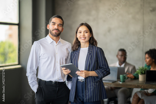 Two professionals stand together in a stylish office, engaging with digital devices. Behind them, a diverse team discusses projects, reflecting teamwork and international cooperation.