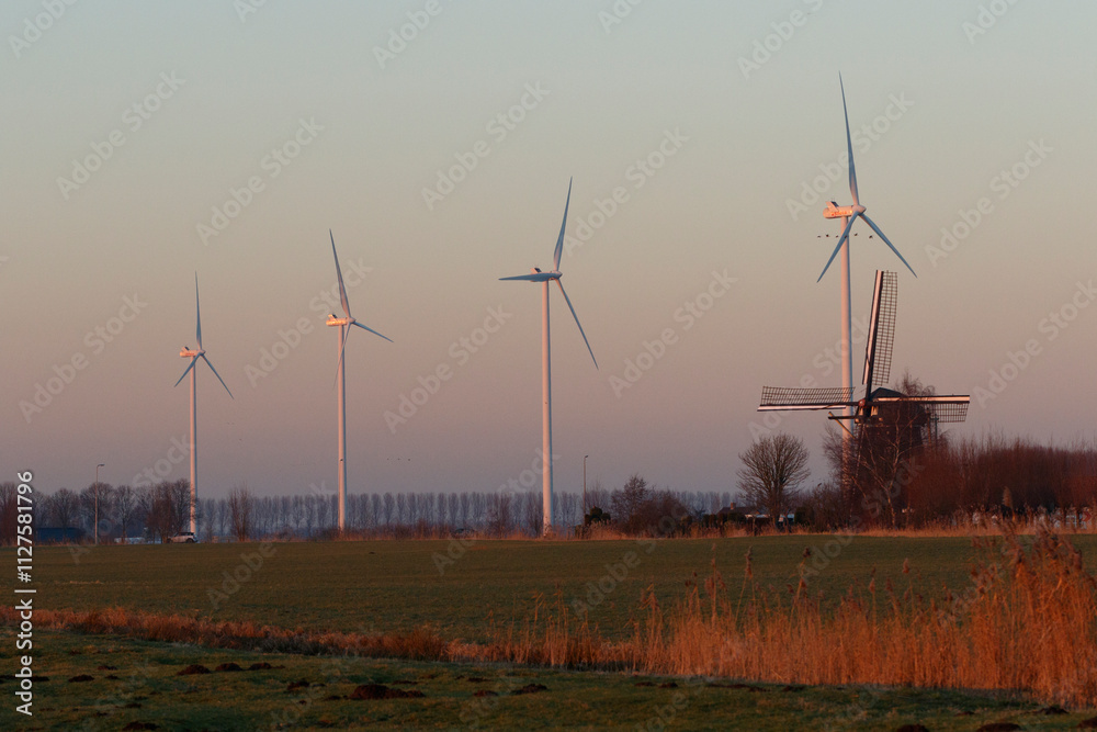 Obraz premium Wind turbines and a traditional windmill stand in a field at dawn or dusk. Rural Dutch landscape.