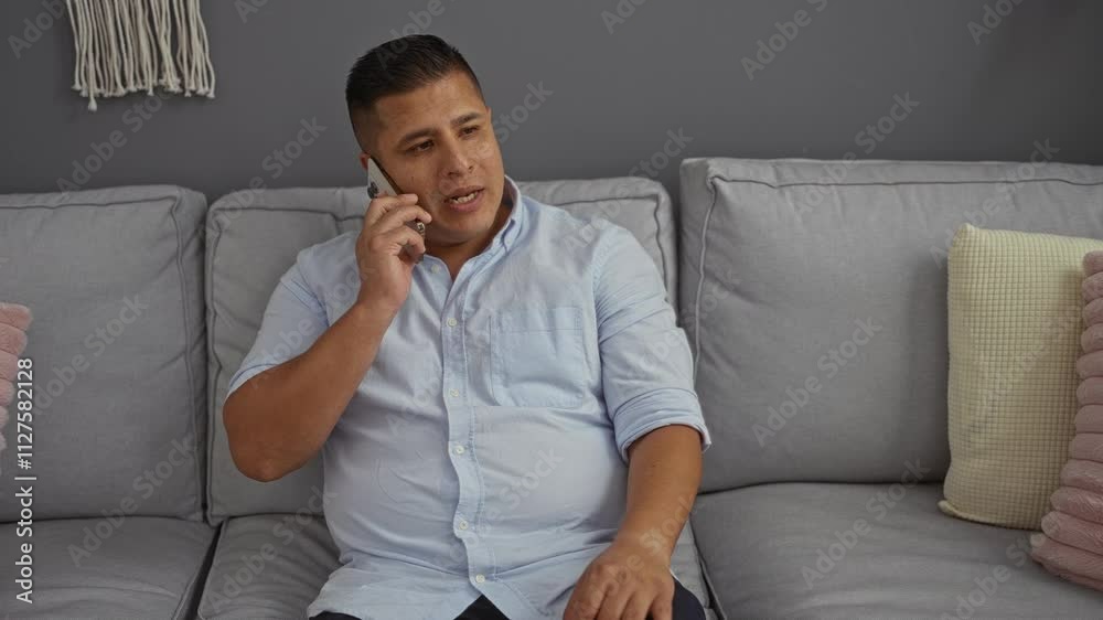 Young man talking on smartphone while sitting on gray couch in living room of his home Young man talking on smartphone while sitting on gray couch in living room of his home