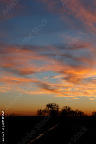 Sunset hues paint a dramatic sky over a dark rural landscape. Silhouetted trees line a waterway.