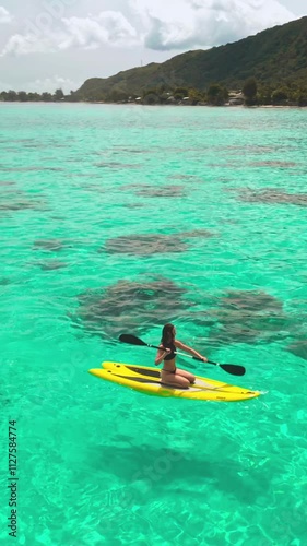 Vertical Screen Video. Aerial drone shot woman paddle boarding coral reef tropical island. Young female rows sitting on yellow paddleboard in crystal turquoise sea water. Outdoor lifestyle travel