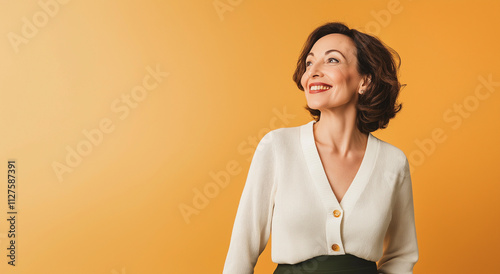 Uma mulher de meia-idade, vestida com um cardigã branco e uma saia verde-escura, sorri levemente com os olhos bem abertos em um estúdio com um fundo laranja suave, irradiando uma felicidade gentil. photo