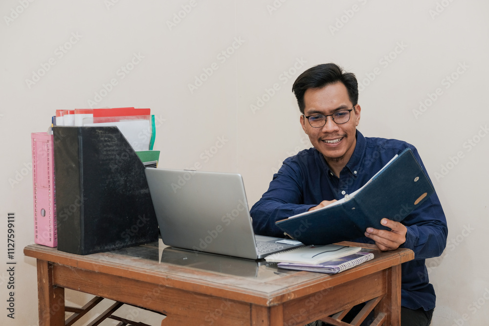 Smiling Southeast Asian Man Working While Sitting In Front Of Laptop and open notepad In Home Office. Workplace Of Professional Workers, Freelancers or Students. Smiling Southeast Asian Man Working While Sitting In Front Of Laptop and open notepad In Home Office. Workplace Of Professional Workers, Freelancers or Students.