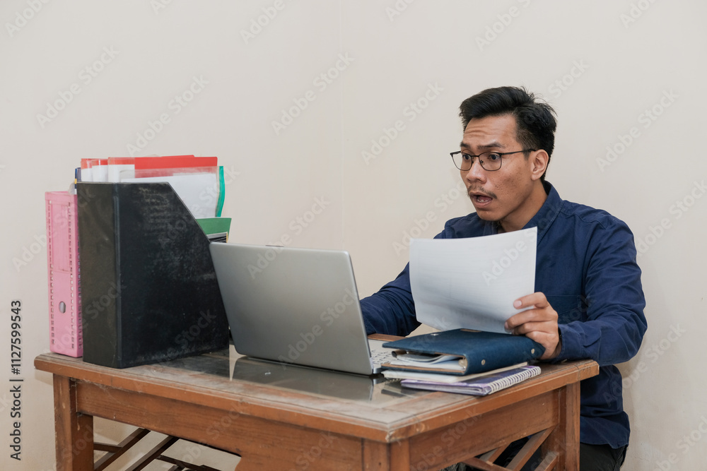 Shocked Southeast Asian Man Works while Sitting in front of a laptop and holding paperworks at Home office. The Workplace of a Professional Worker, Freelancer or Student. Distance Work Concept. Shocked Southeast Asian Man Works while Sitting in front of a laptop and holding paperworks at Home office. The Workplace of a Professional Worker, Freelancer or Student. Distance Work Concept.