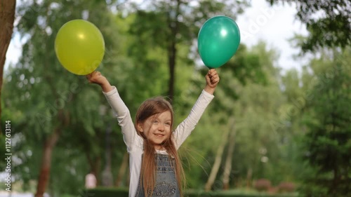 A cheerful and joyful girl is holding a bunch of colorful green and yellow balloons in a vibrant park setting