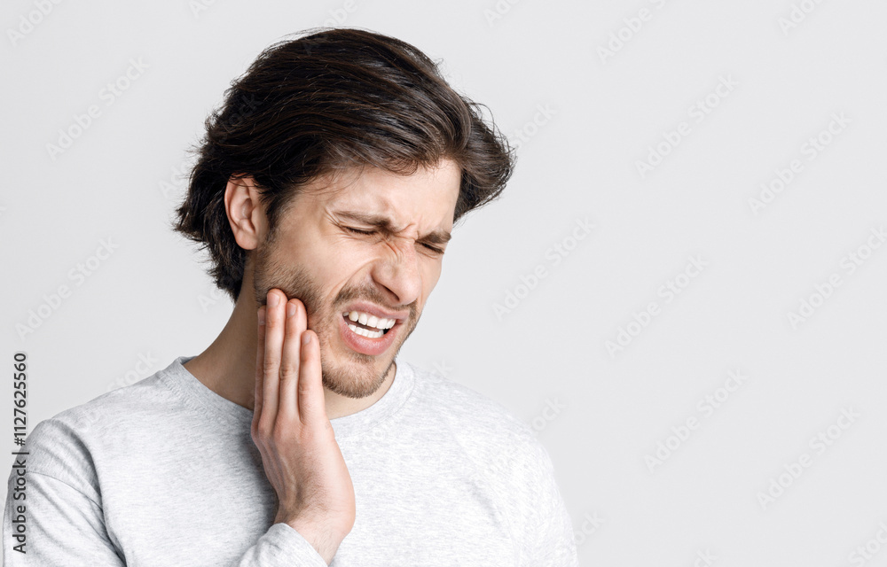 Tooth disease. Man feels pain, grimaces and press hand to cheek isolated on gray background, studio shot