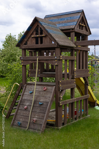 Wooden children's playground with climbing wall, rope, and slide in a backyard garden on a cloudy day, promoting active outdoor play, creativity and physical development for kids in a safe environment