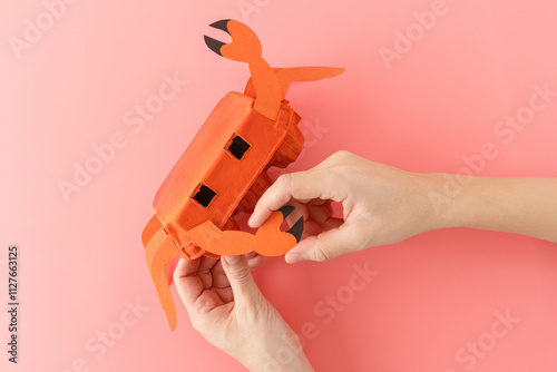 top down view of hands crafting DIY crab toy made from orange painted egg carton and paper cutouts. Step-by-step collage on a pink background. Craft project for kids, photo