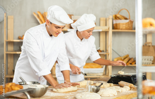 Two young bakers prepare dough in kitchen of bakery - together they knead dough, cut it with a knife and weigh it on scales photo