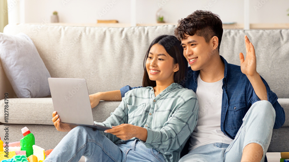 Cheerful asian couple with cleaning tools sitting on floor in living room, having video call, using computer, looking for house-keeping service on Internet. Tired young man and woman with notebook Cheerful asian couple with cleaning tools sitting on floor in living room, having video call, using computer, looking for house-keeping service on Internet. Tired young man and woman with notebook