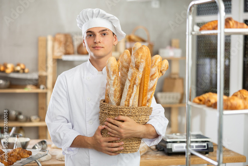 Positive young guy wearing pinafore holding baguettes in round wicker basket in bakery shop photo