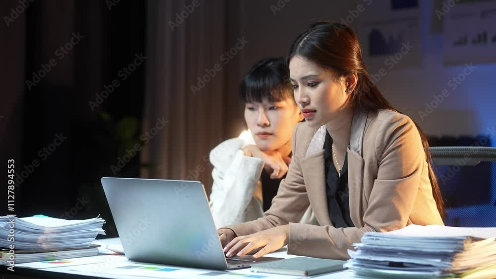 Two women are sitting at a desk with a laptop open in front of them. They are looking at the screen and seem to be working on something together Two women are sitting at a desk with a laptop open in front of them. They are looking at the screen and seem to be working on something together