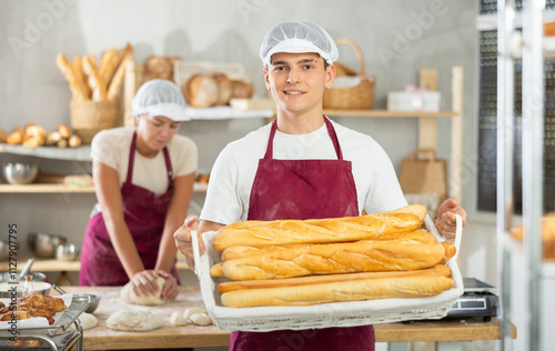 Positive young guy wearing pinafore holding baguettes in square wicker basket in bakery shop photo