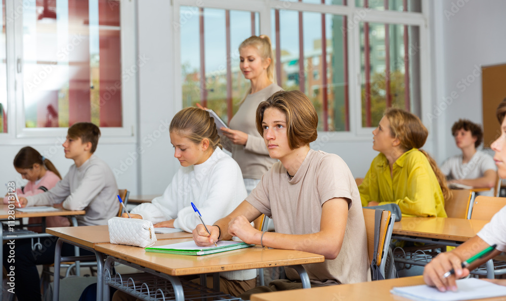 Teen boys and girls sitting at desk in classroom full of pupils during lesson Teen boys and girls sitting at desk in classroom full of pupils during lesson