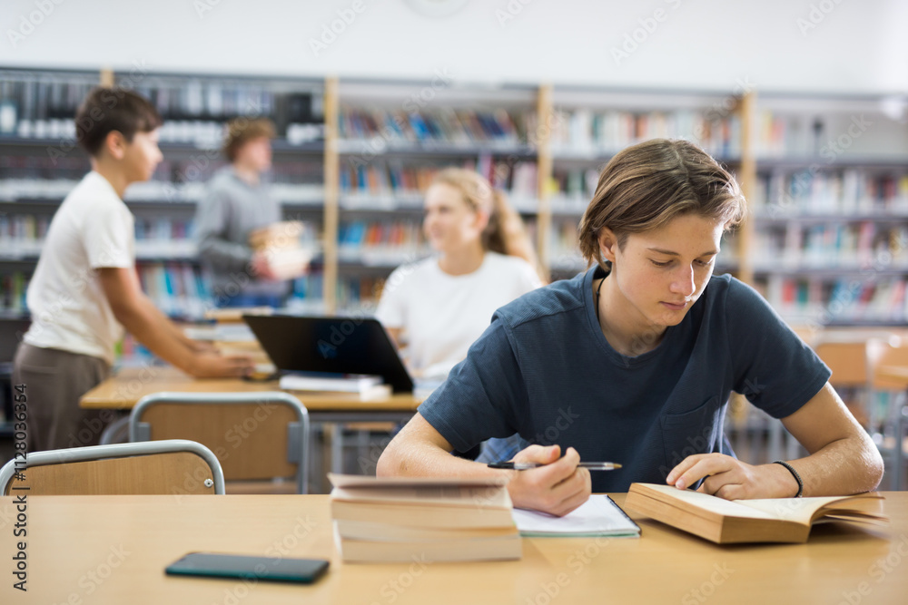 Fifteen-year-old schoolboy studying in the library writes an synopsis from a textbook Fifteen-year-old schoolboy studying in the library writes an synopsis from a textbook