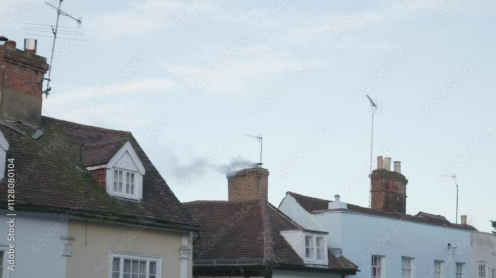Rooftops of older houses, each a different colour—baby blue, yellow, and white. The central house is notably shorter, with smoke rising from its chimney against a clear sky Rooftops of older houses, each a different colour—baby blue, yellow, and white. The central house is notably shorter, with smoke rising from its chimney against a clear sky