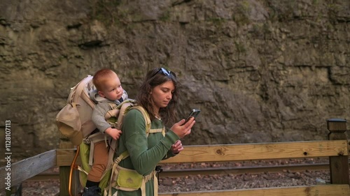 Mother with her child in backpack pauses to check her phone along wooden walkway near cliffs at Natural Tunnel State Park in Virginia, USA. Mother with her child in backpack check her phone photo