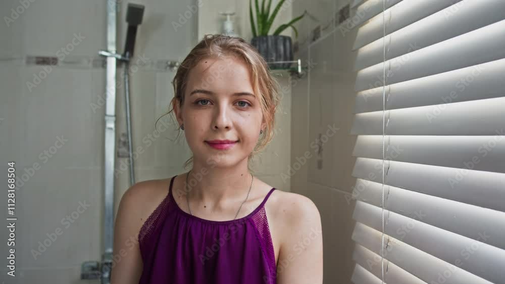 Medium portrait of young Caucasian woman standing by bathroom window with blinds, softly lit by natural light Medium portrait of young Caucasian woman standing by bathroom window with blinds, softly lit by natural light