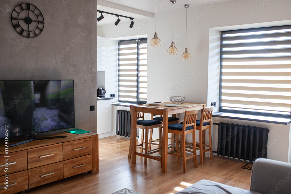 Fototapeta premium Modern open kitchen interior with wooden high bar table and chairs, sofa and TV, blinds on windows. Stylish living room with dining space. Selective focus.
