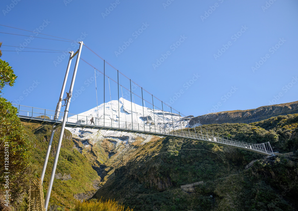 Tourist walking on Manganui Gorge suspension bridge. Snow-capped Mt Taranaki in the background. Taranaki. New Zealand.