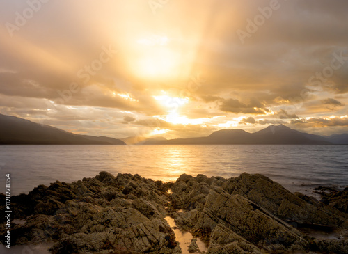 Fototapeta Naklejka Na Ścianę i Meble -  Sunrise over water by a mountain and sea landscape with golden sun beams shining through low clouds, with rocks in the foreground, shot at Duisdealmor on Isle of Skye, Scotland