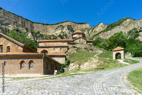 A paved road leads past the refectory to the church of the Shio-mgvime monastery. A rock with cave cells is in the background. photo