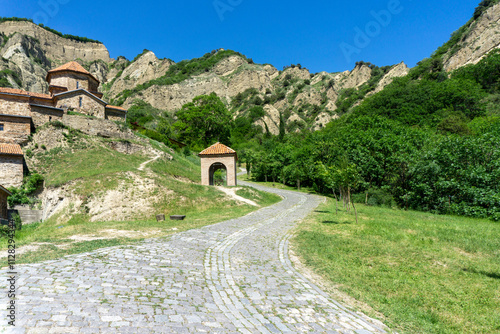 A paved road leads past the refectory to the church of the Shio-mgvime monastery. A rock with cave cells is in the background. photo