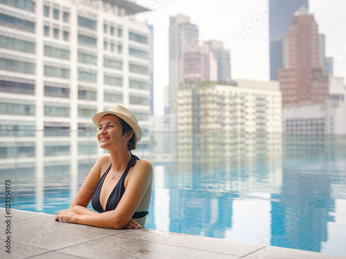 Young asian woman relaxing by pool at Kuala Lumpur hotel with view of surrounding skyscrapers, enjoying leisure time in vibrant urban setting.