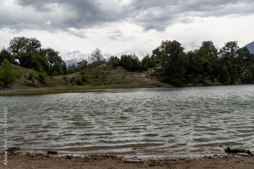 A serene lake with gentle ripples reflects the cloudy sky, surrounded by dense green trees and distant snow-capped mountains. Peaceful overcast atmosphere at Deoria tal lake trek in Ukhimath, India. A serene lake with gentle ripples reflects the cloudy sky, surrounded by dense green trees and distant snow-capped mountains. Peaceful overcast atmosphere at Deoria tal lake trek in Ukhimath, India.