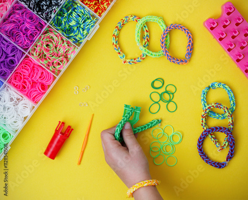 Child's hand holding a work in progress loom bracelet, other colorful elastic bracelets and bracelet making supplies on yellow background. Toy developing children's imagination. Flat lay, from above.