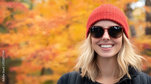 A blonde woman wearing sunglasses and red beanie smiling in autumn, with fall foliage background