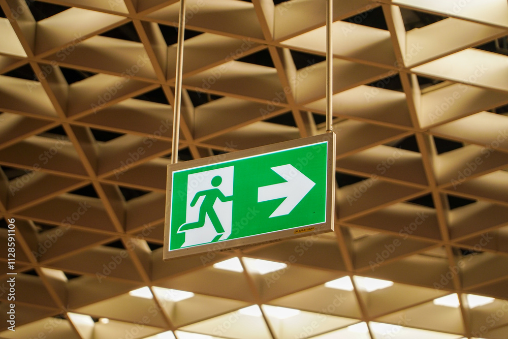 A green emergency exit sign hangs from a geometric ceiling. The sign shows an arrow and running figure indicating the direction to an emergency exit. A green emergency exit sign hangs from a geometric ceiling. The sign shows an arrow and running figure indicating the direction to an emergency exit.