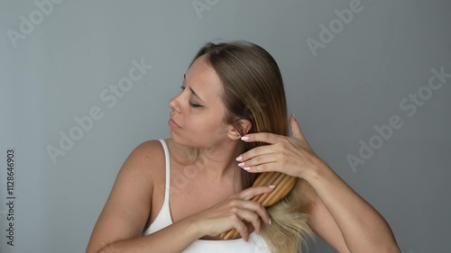 Young caucasian blonde woman combing her dry straight hair with a wooden comb on a dark grey color background photo