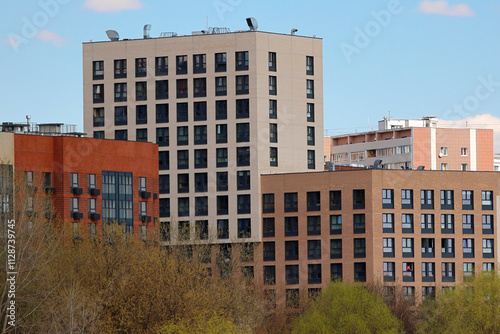 A Cluster Of Contemporary Apartment Buildings Under A Clear Blue Sky. The Structures Showcase Diverse Architectural Styles, With Leafy Trees In The Foreground Adding A Touch Of Nature.