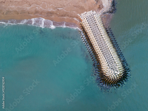 Fototapeta Naklejka Na Ścianę i Meble -  High-angle view of a coastal breakwater extending into turquoise water.This aerial shot showcases a breakwater, a series of large concrete blocks extending into the turquoise ocean, creating a distinc