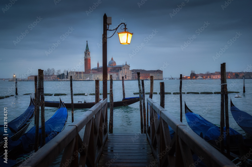 Evening view of gondolas at a pier with Venetian architecture illuminated by a street lamp Evening view of gondolas at a pier with Venetian architecture illuminated by a street lamp