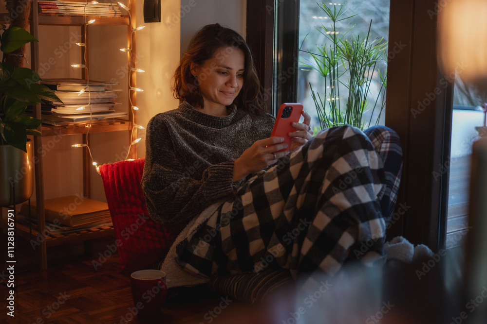 Young Caucasian woman sitting comfortably at cozy home with smartphone during winter Christmas holidays