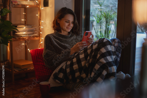 Young Caucasian woman sitting comfortably at cozy home with smartphone during winter Christmas holidays photo