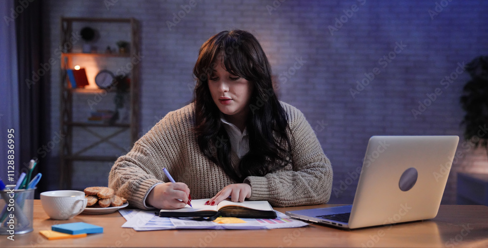 Young business woman freelancer sitting at desk working on remote in home office late, using laptop and writing notes in notebook, focused expression.