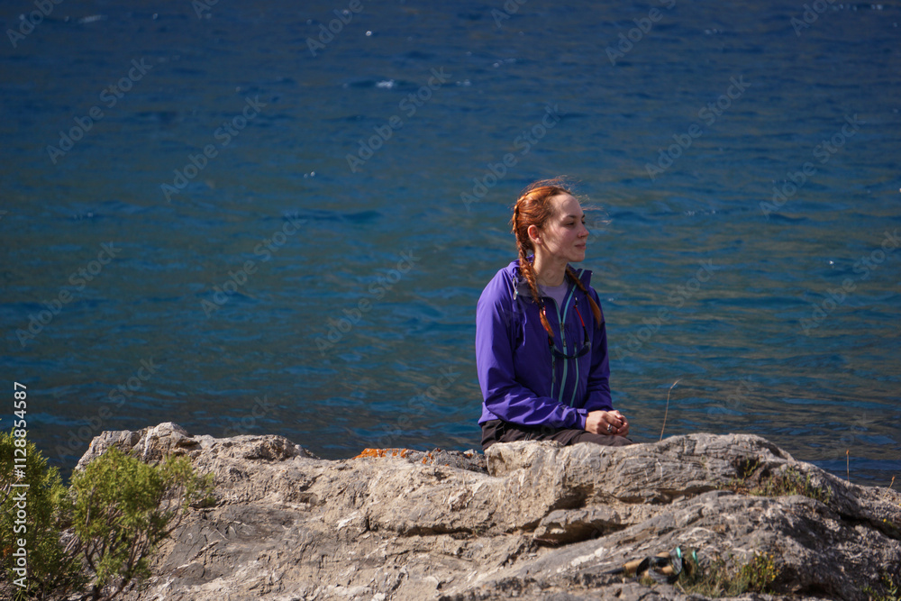 Fototapeta premium A young girl sits on a rock in the middle of a blue mountain lake