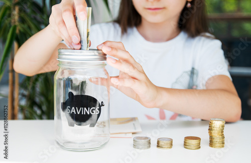 Cute girl puts banknotes in a piggy bank, stacks of coins on the table.