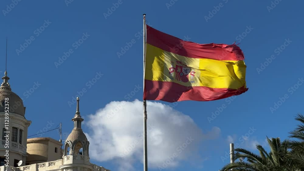 Spanish flag waving against blue sky with architectural domes and palm trees in Alicante, Valencia, Spain