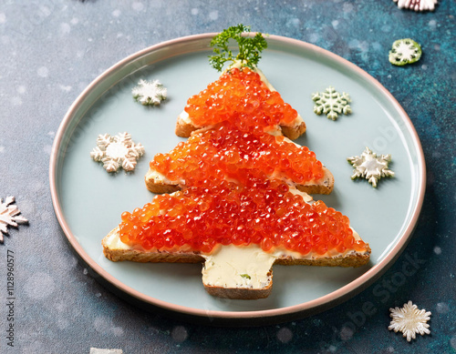 A festive Christmas tree-shaped toast is generously topped with bright red caviar and garnished with fresh herbs, served on a light blue plate decorated with snowflake ornaments. photo