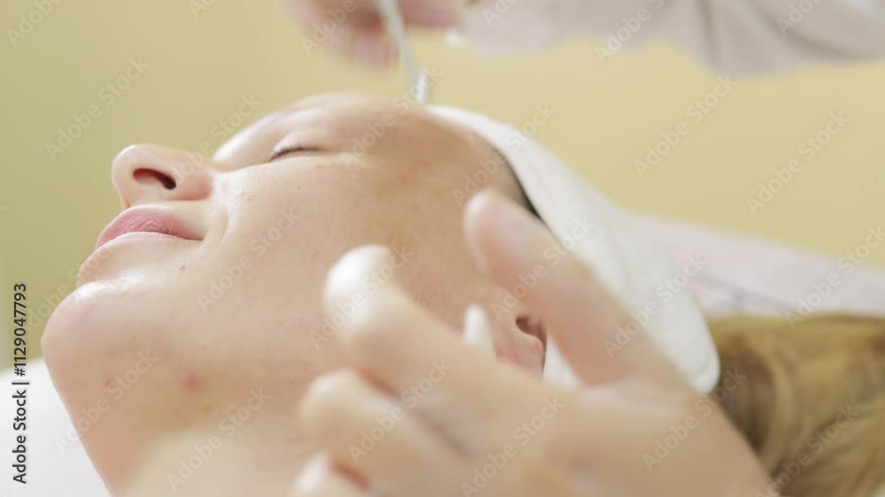 Beautician wearing latex gloves removes impurities from woman's face using metal tool during professional treatment. Beauty Clinic Concept Beautician wearing latex gloves removes impurities from woman's face using metal tool during professional treatment. Beauty Clinic Concept