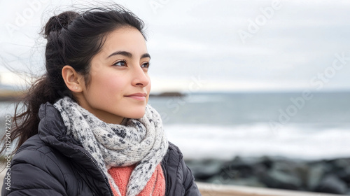 Hispanic woman sitting on a bench by the ocean, wearing a coat and a scarf