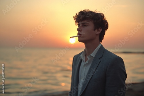 As the sun sets, a young man in a grey suit stands on the beach, cigarette in mouth, watching the last rays disappear 2 photo