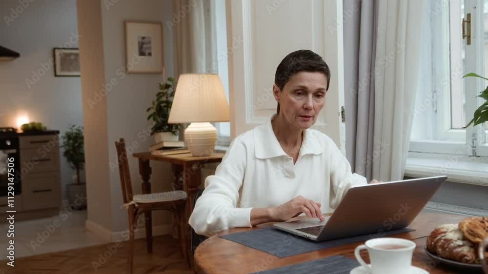 Senior elegant woman sits at table typing on keyboard. Cheerful aged woman working remotely using laptop. Older focused businesswoman browsing online. Female elegant writer enjoying creative process